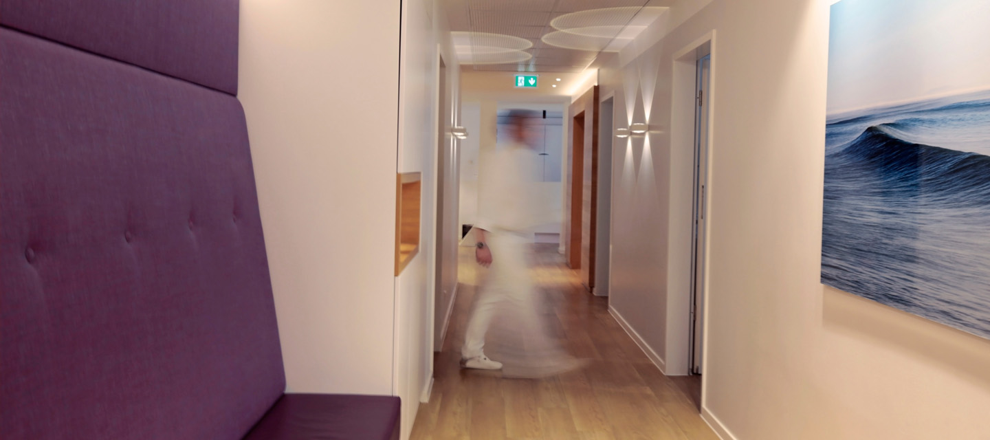 Moderner Wartebereich vor den Praxisräumen mit einem violetten Sofa. Im Hintergrund geht eine Person durch den Flur; die Bewegung ist leicht verschwommen dargestellt.